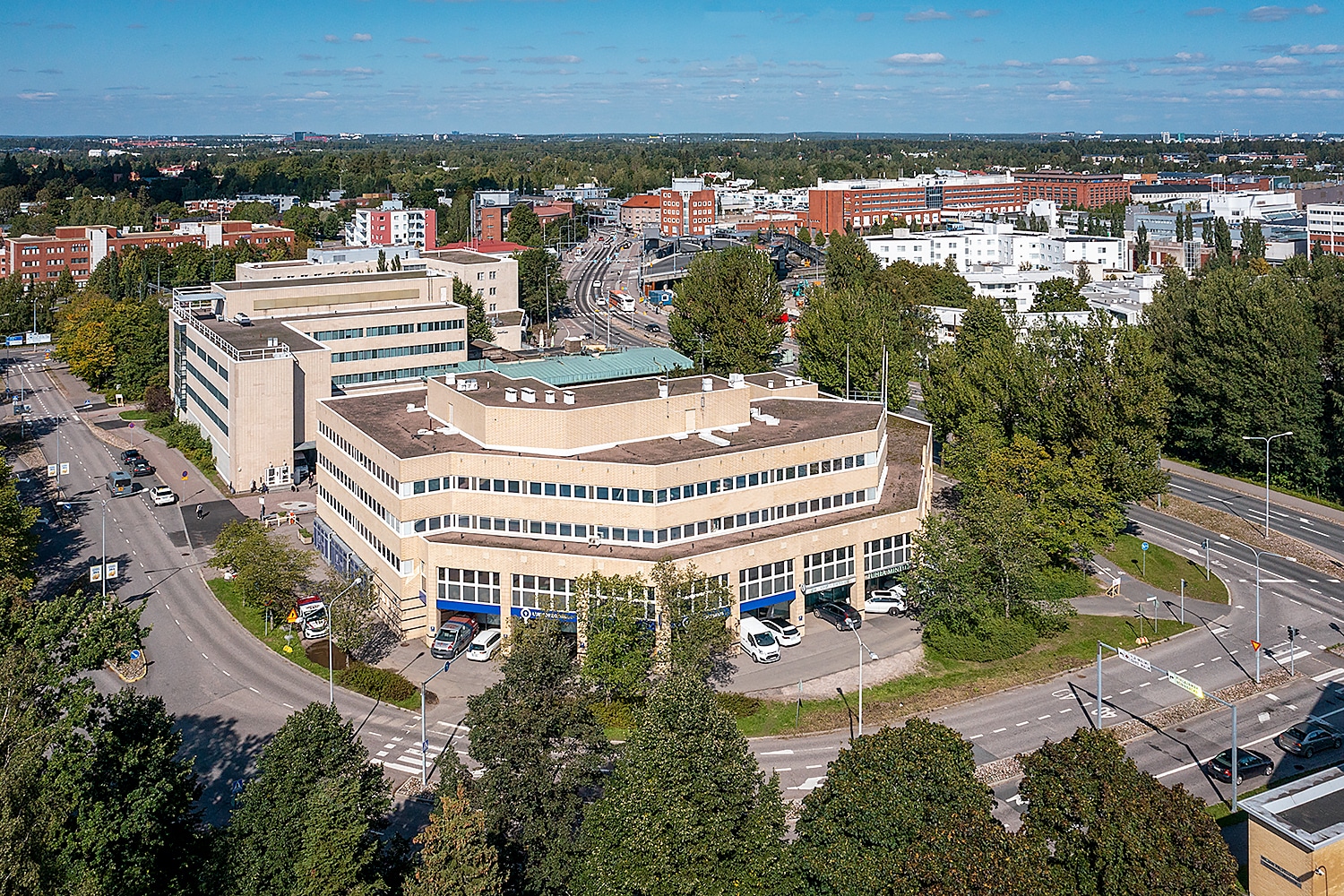 Ilmakuvassa u:n muotoinen toimitilakiinteistö Vanha Helsingintie 18. Ympäri talon kellertävät seinät ja nauhaikkunat. Taustalla näkyy Malmin keskustaa. Talon vierellä puita.