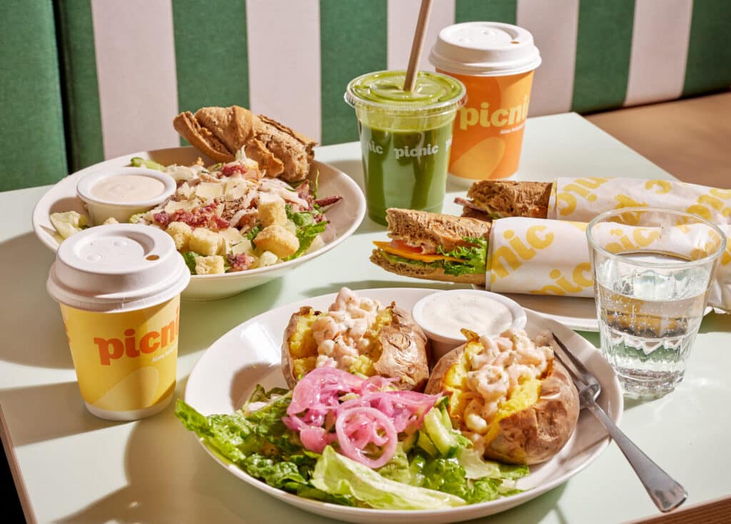 Kaksi Picnicin uuniperuna-annosta, smoothie, kaksi täytettyä patonkia ja kaksi kahvia pöydällä.